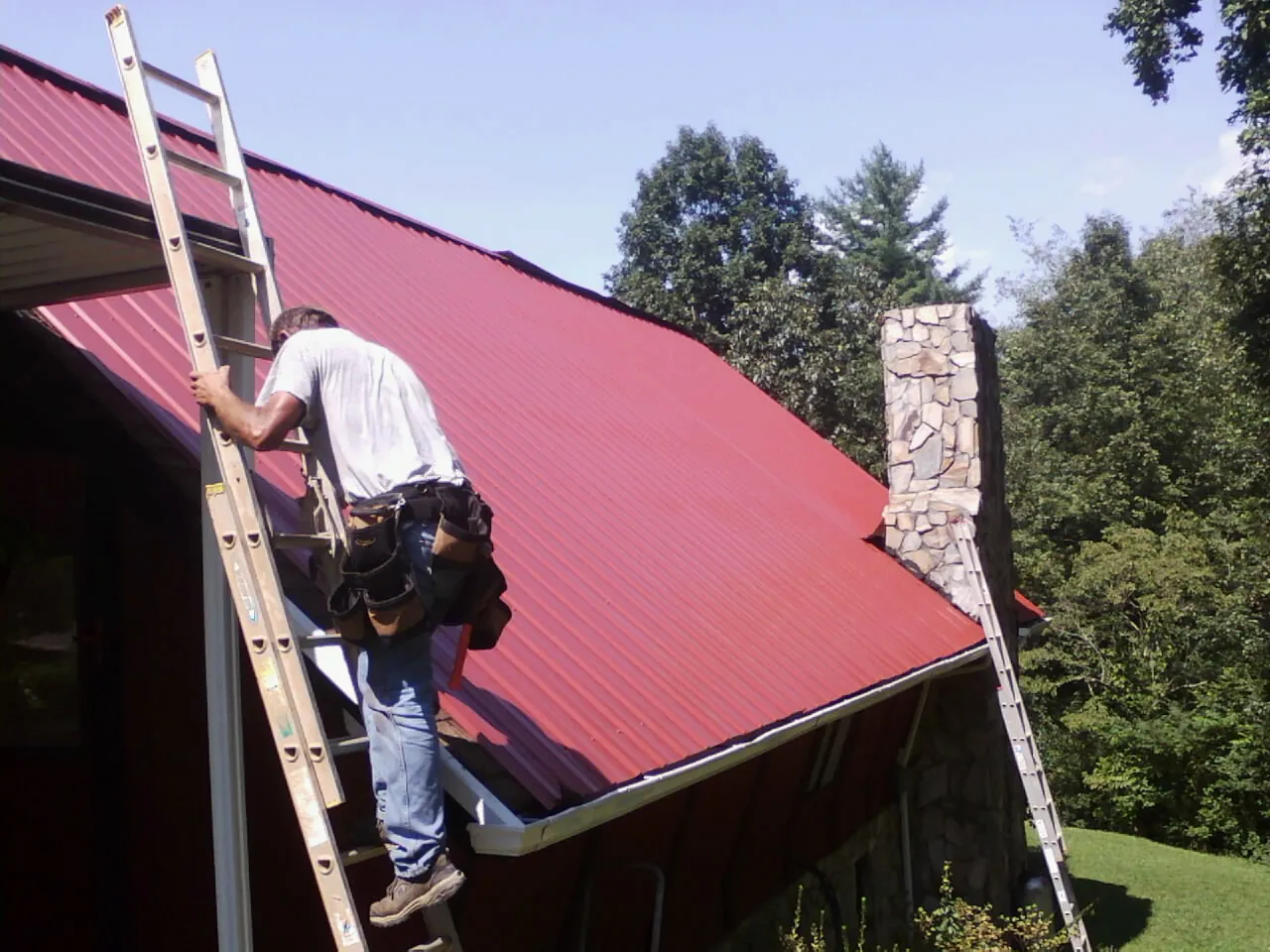 Roofer on a ladder beside a red metal roof and stone chimney — Soffit & Fascia Repair by Roofing Brothers in Arnaudville